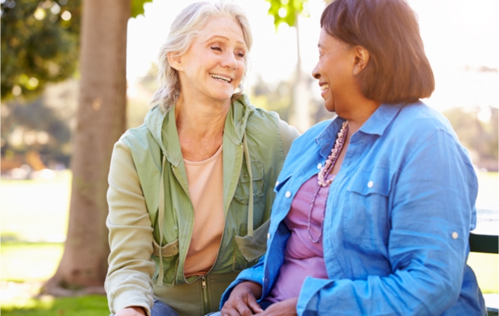 Two mature women sitting and laughing on a bench surrounded by trees