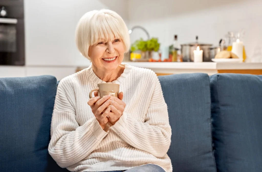 A smiling mature resident sits on a blue couch while enjoying a cup of tea.
