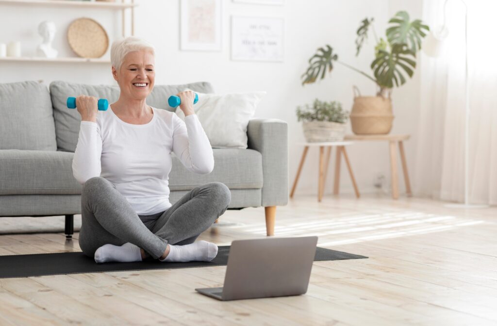 An older smiling resident sitting cross legged on a yoga mat as she holds up two small dumbbells next to her shoulders.