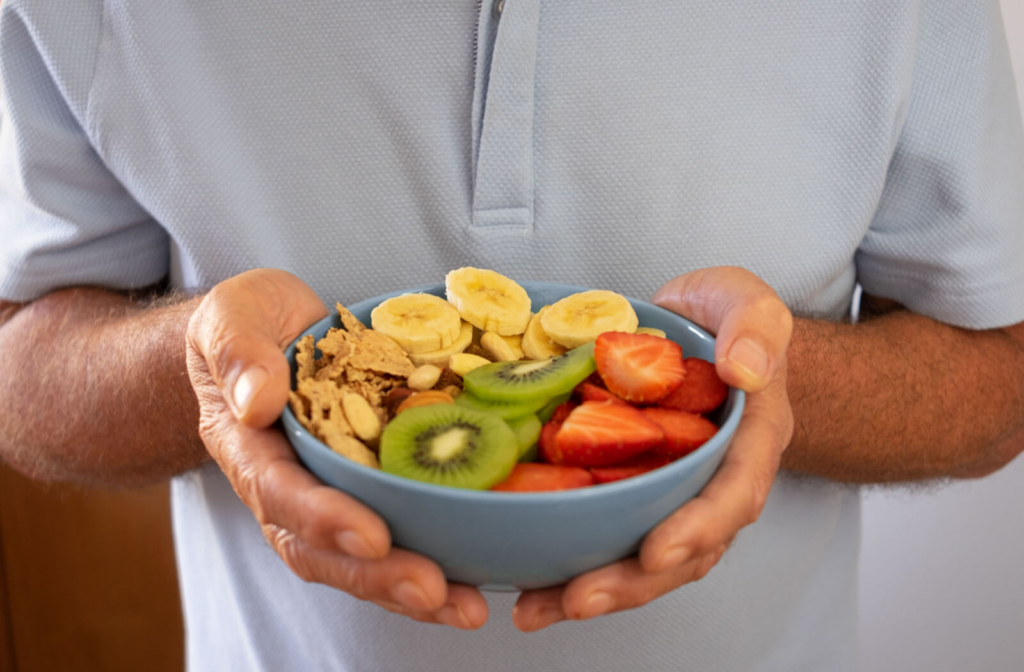 A senior man holding a bowl with granola, kiwi, bananas, and strawberries.