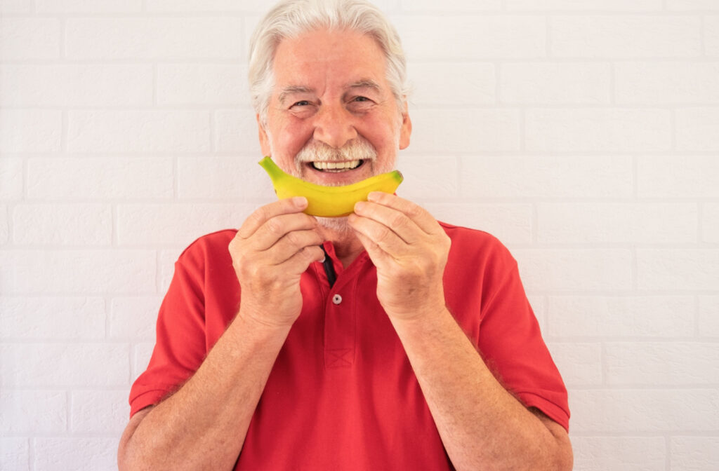 A senior man smiling with a white beard and in red polo shirt, holding a banana in front of his mouth like a smile.