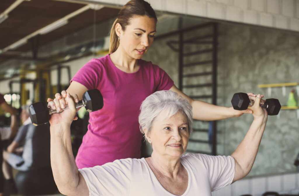 An older adult woman, who is lifting a pair of light weight dumbbells, being assisted by a fitness coach.