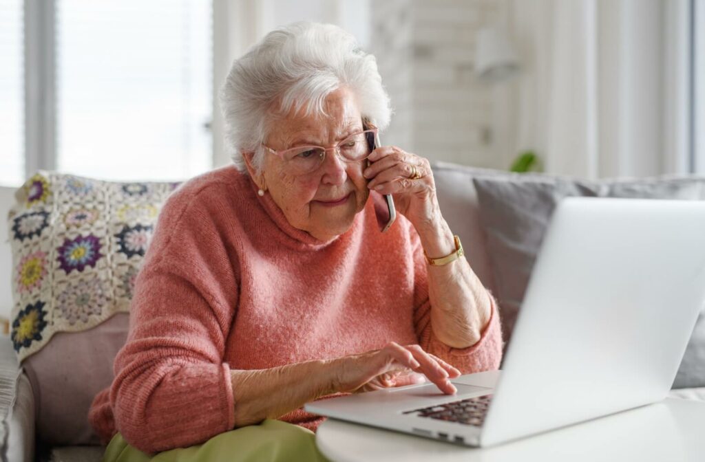 An older adult making a phone call while using their laptop to make sure a website is safe.