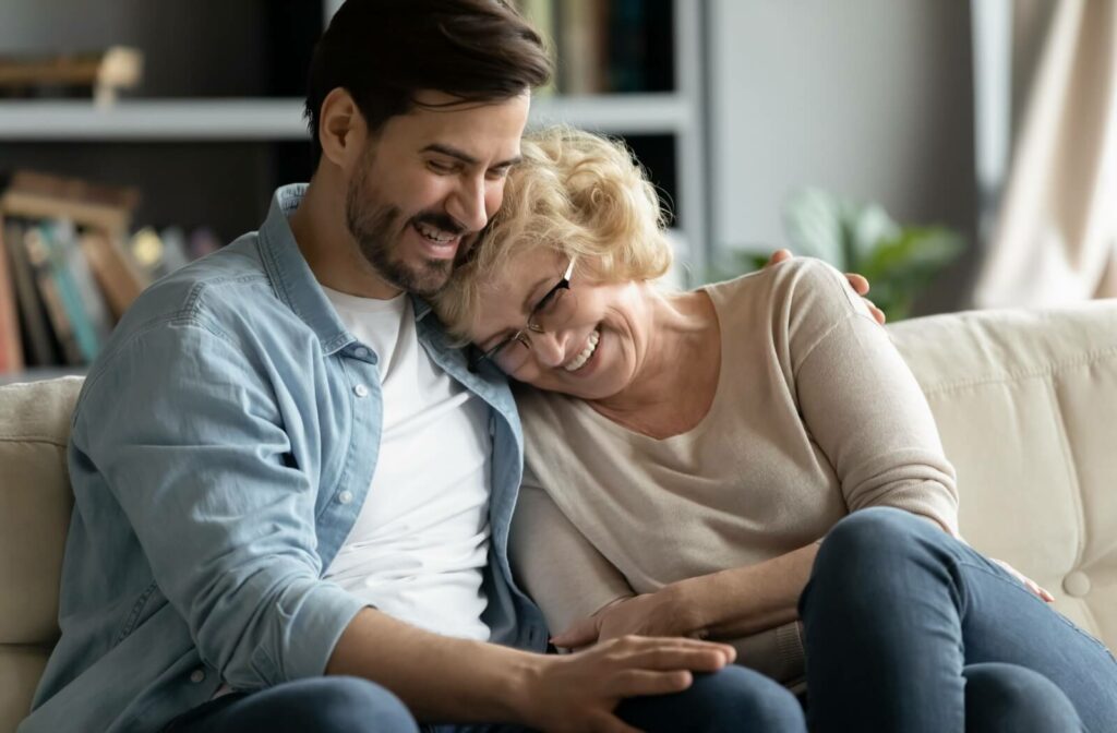 An adult laughing and hugging their older parent on the couch during a visit.