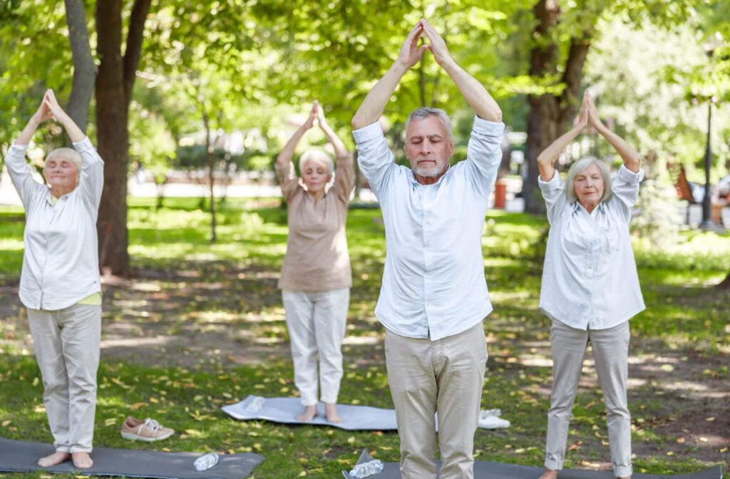 Standing in a park, 4 older adults stand in a tower pose during a meditative Tai Chi class to promote relaxation