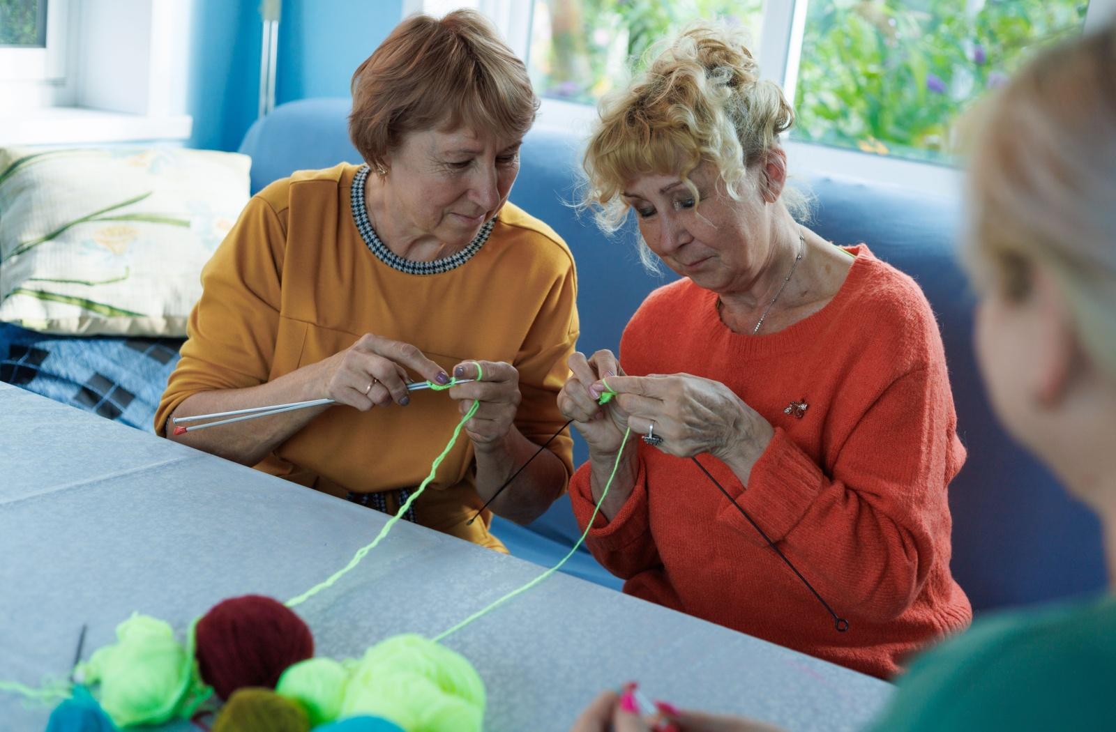  Two older adults smile while knitting together during an arts class in independent living