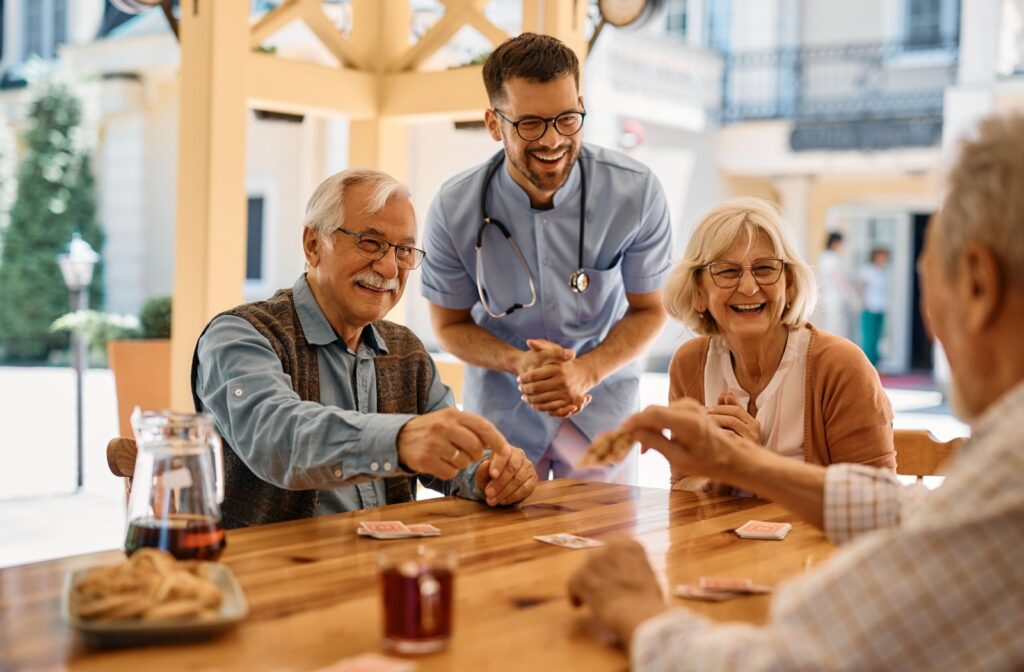 A group of senior friends smile as they enjoy playing cards while a smiling caregiver watches.
