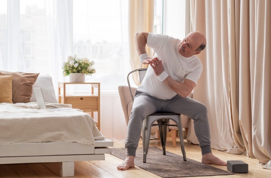 A senior sits on a chair and performs a light arm stretch.