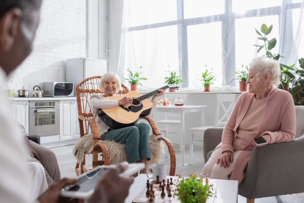Happy senior woman playing acoustic guitar near multiethnic friends with devices and chess in nursing home